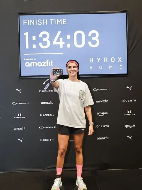 Nikita Kohli of Equilateral Training, functional fitness facilitator for the Active Day at EkkoFarm, Bétera, Valencia, in training kit holding up a HYROX Rome card in front of a finish-time screen